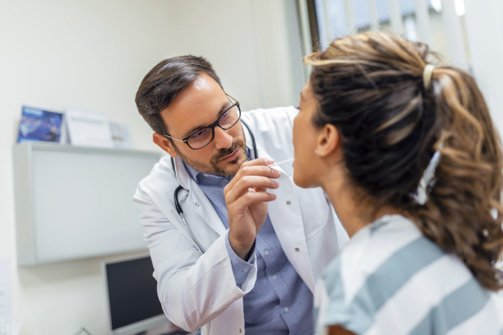 A young woman sits on an exam table across from her doctor. The doctor reaches forward with a tongue depressor as the woman looks up and sticks out her tongue.